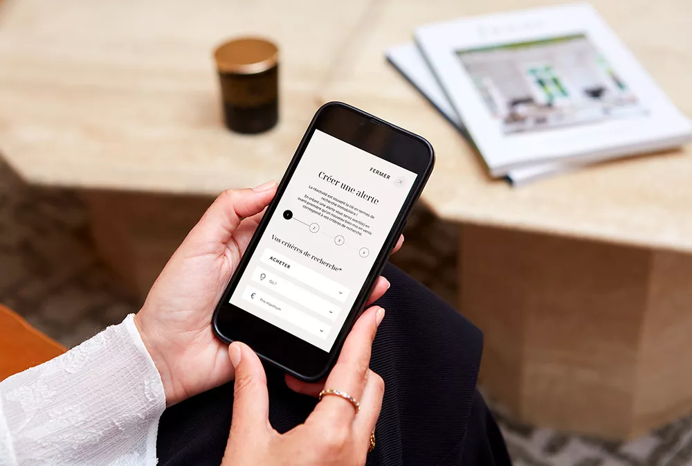 Un client Junot d'une entreprise tient un téléphone ouvert sur le site internet Junot et crée une alerte de nouveau bien