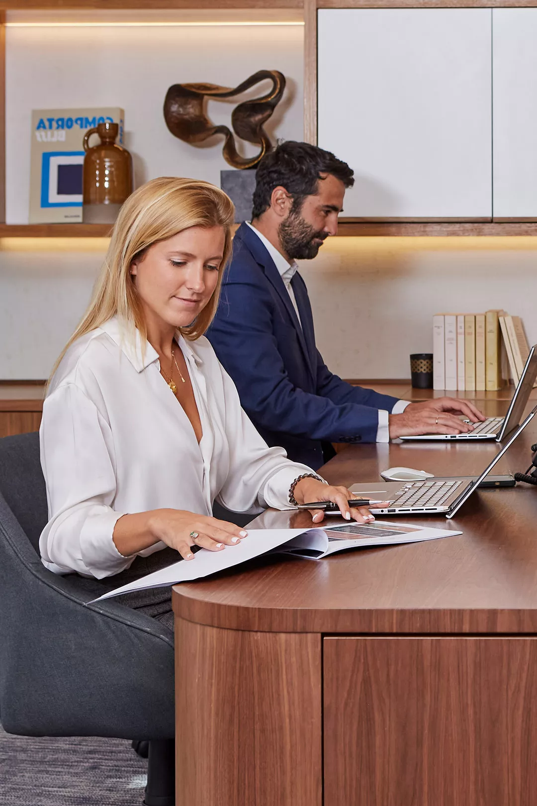 A man in a blue suit writing on a piece of paper and a woman on the phone in the background